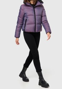 Chaqueta de plumas en púrpura iridiscente, con cierre frontal, bolsillos laterales y detalles en negro. Combinada con pantalones negros y botas negras gruesas.