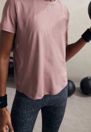 Osoba ubrana w różowy T-shirt marki Nike i wzorzyste legginsy trzyma czarną piłkę lekarską, obok na podłodze znajdują się dodatkowe piłki lekarskie.