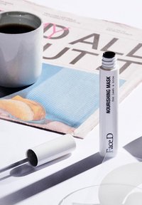 Un tubo bianco etichettato "MASCHERA NUTRIENTE" con cappuccio nero, circondato da una tazza bianca, un bicchiere e un giornale a motivo rosa e blu.