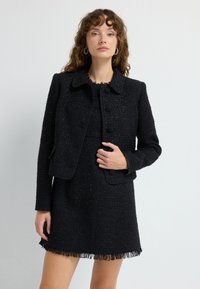 Sort tweedjakke med krave, frontknapper og lommer. Kombineret med en matchende kjole med frynsekant, begge i tekstureret stof.
