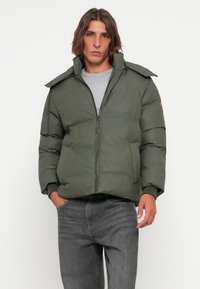 Puffer jacket in dark green, featuring a full-length zipper, wide collar, and ribbed cuffs. Worn over a gray shirt with dark jeans.