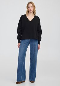 Zwarte gebreide trui met een V-hals en drop schouders, gecombineerd met lichtblauwe, hoge taille, wijd uitlopende jeans en schoenen met dierenprint.