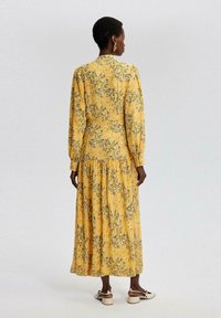Maxime dress in keltakanga, joka esittelee kukkakuviota sinisessä ja harmaassa. Pitkät hihat, kerroksellinen helma ja korkea kaula-aukko.