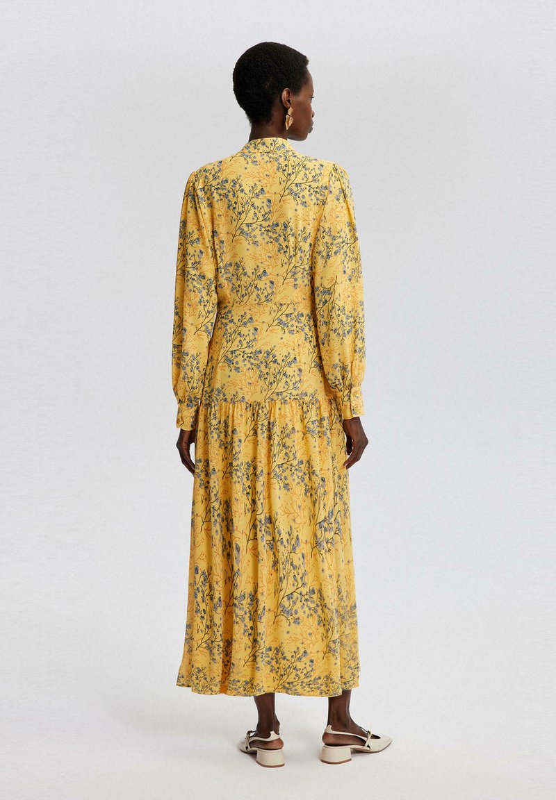 Maxikleid aus gelbem Stoff mit floralem Muster in Blau und Grau. Lange Ärmel, ein gestuftes Saumdesign und ein hochgeschnittener Kragen.