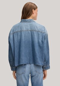 Denim jacket in light blue with a cropped cut, long sleeves, and button cuffs. Features a yoke seam and frayed hem detail.