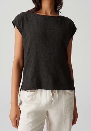 Kobieta w teksturowanym czarnym topie z krótkim rękawem w połączeniu z luźnymi białymi spodniami ze ściągaczem na jednolitym tle.