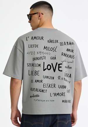 Uomo che indossa una maglietta grigia con la parola "love" scritta in varie lingue sulla schiena, visto da dietro.