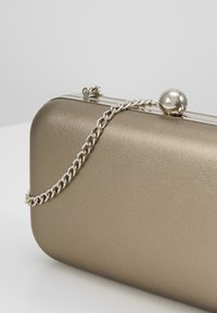 Pochette rectangulaire en métal doré avec une bandoulière chaîne argentée polie et un fermoir rond argenté sur le dessus.