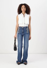 Λευκή κοντή αμάνικη μπλούζα με υφή, ψηλόμεσα μπλε τζιν με χρυσά κουμπιά, μαύρη τσάντα χειρός και ψηλοτάκουνες γόβες.