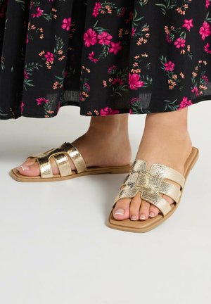 Fødder iført gyldne metalliske flade sandaler med udskårne stropper, kombineret med et sort nederdel med pink og orange blomsterprint.