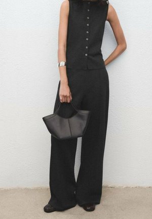 Femme portant un top noir sans manches boutonné et un pantalon à jambes larges tenant un sac à main noir contre un mur blanc.