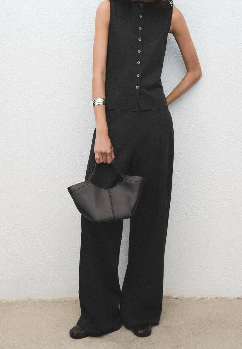 Femme portant un top noir sans manches boutonné et un pantalon à jambes larges tenant un sac à main noir contre un mur blanc.