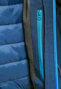 Section d'une veste doudoune bleue avec une texture lisse, présentant un panneau en tissu gris foncé et une poche zippée turquoise vif.