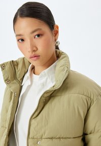 Giacca a piumino verde oliva con collo alto, accessori in argento e cuciture visibili. Indossata sopra una camicia bianca. Texture liscia.