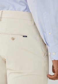 Béžové bavlněné kalhoty s knoflíkovou zadní kapsou a modrou nášivkou, v kombinaci se světle modrou košilí s detaily knoflíků.
