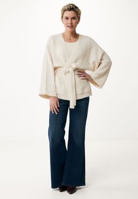 Krämfärgad cardigan med breda ärmar och knytbälte, matchad med mörkblå utsvängda jeans och vinröda skor. Minimalistisk design och neutrala färger.