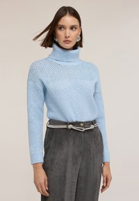 Ljusblå stickad tröja med hög krage, med ett nätmönster, ihop med mörkgrå kordverksbyxor med ett dekorativt bälte.