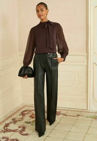 Tamno smeđa bluzica s mašnom oko vrata i prozirnim balon rukavima, uparenim s visokim strukom, naboranim crnim kožnim hlačama i crnom clutch torbom.