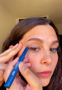 Blauwe eyelinerpotlood vastgehouden door een persoon die het op het ooglid aanbrengt. Het potlood is cilindrisch met een matte afwerking en is van gemiddelde lengte.