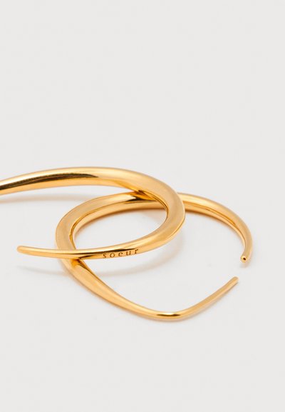 Dwie skręcone złote kolczyki koła z gładkim wykończeniem, jeden z wygrawerowanym słowem "soeur", prezentowane na białym tle.