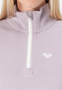 Light purple fleece pullover with white zipper and small heart-shaped logo on the left chest, worn by a person with blonde hair.
