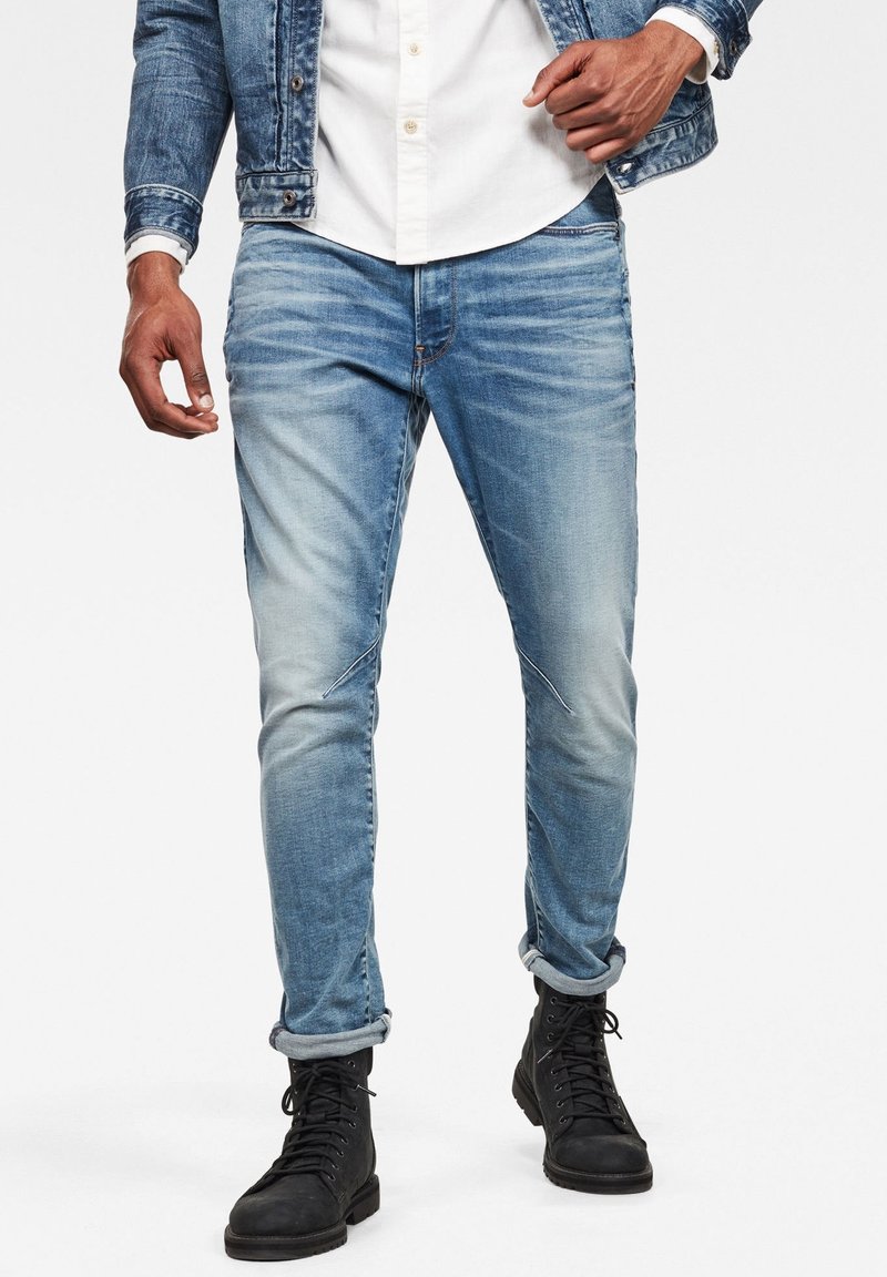 Světle modré slim fit džíny z denimu s vybledlým efektem. Mají vyhrnuté lemy a složité prošití podél nohou, doplněné černými botami.