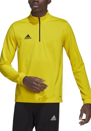 Gele sportieve top met kwart ritssluiting, gemaakt van gladde stof, met een zwart adidas-logo en aansluitende manchetten, gecombineerd met zwarte broek.