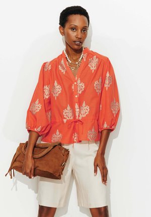 Femme portant un chemisier orange à motifs et un short blanc, tenant un sac à main en daim marron avec des détails dorés, posant sur un fond blanc.