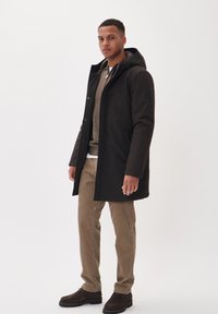 Black, knee-length parka with hood, front zip and snap button closure. Worn over a beige sweater and tan corduroy pants, paired with dark shoes.