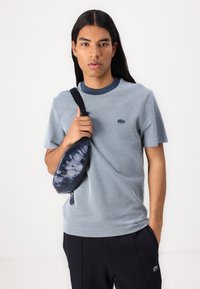 Mėlyna trumpomis rankovėmis marškinėliai su kontrastinga tamsia apykakle ir Lacoste logotipu. Ant peties nešiojama tamsiai mėlyna tie-dye stiliaus per petį dėvima rankinė.