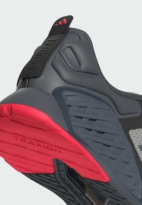 Szare buty sportowe z teksturowaną cholewką, czarnymi akcentami i czerwoną podeszwą antypoślizgową. Posiadają wyraźny obcas i wzory poprawiające przyczepność.