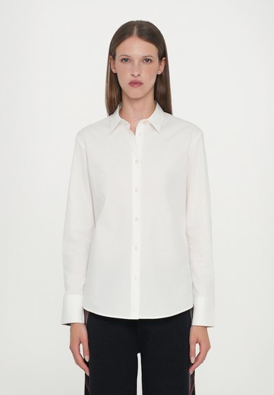 Chemise blanche à manches longues avec un col pointu, fermeture à boutons sur le devant et coupe droite. Le tissu semble lisse et léger.