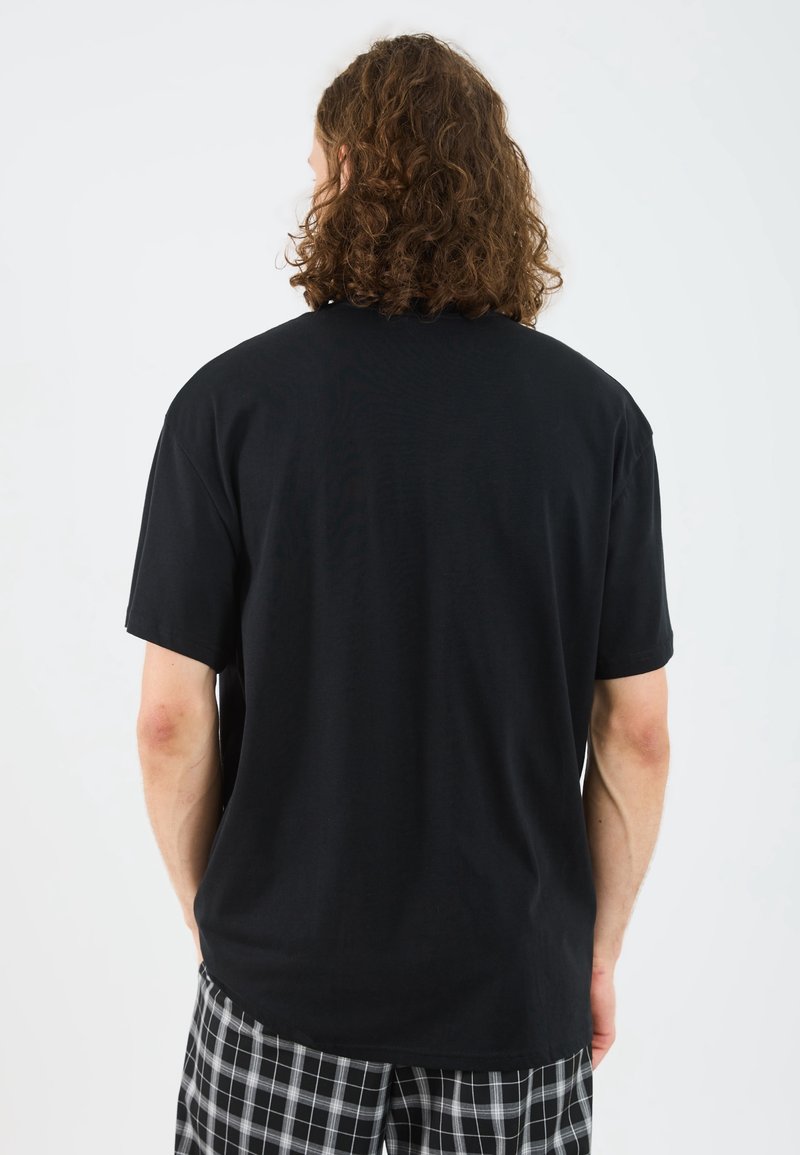 Personne aux cheveux bruns bouclés, portant un t-shirt noir uni à manches courtes et un pantalon à carreaux noir et blanc, debout dos à la caméra.