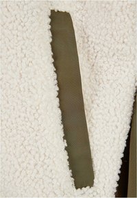 Veste en polaire avec une texture blanche duveteuse, agrémentée d'un accent en tissu vert olive intégré dans le design.