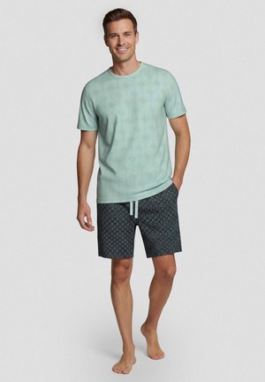 Mann lächelt, trägt ein hellgrünes kurzärmeliges Hemd mit Muster und dunkle gemusterte Shorts, steht barfuß vor einfarbigem Hintergrund.
