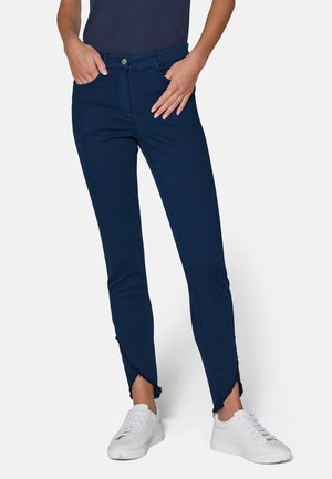 Jean skinny bleu marine avec un ourlet effiloché et angulaire, fabriqué dans un tissu extensible, associé à des baskets blanches. Poches avant incluses.