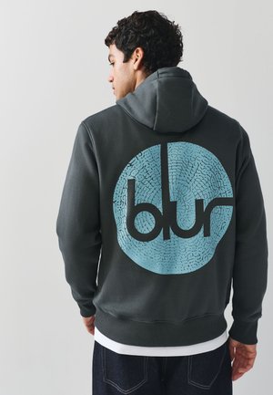 Mann, seitlich stehend, trägt einen dunklen Kapuzenpullover mit großem blauem kreisförmigem „blur“-Logo auf dem Rücken, kombiniert mit dunklen Jeans.