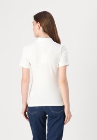 Valge lühikese varrukaga polo, millel on krae, sile tekstuur ja sobiv disain, kombineeritud tumedate siniste teksadega ja ühtlase taustaga.