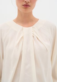 Blusa crema con scollatura rotonda e dettaglio plissettato sul davanti. Realizzata in tessuto leggero e liscio; presenta una sottile lucentezza e una texture morbida.