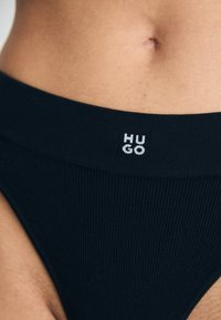 Musta ribbed bikinit laajalla vyötärönauhala, jossa on valkoinen "HUGO"-logo. Sujuva pinta ja minimalistinen muotoilu.