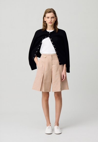 Cárdigan negro con botones dorados sobre una camiseta blanca, acompañado de pantalones cortos de talle alto a rayas beige y naranja. Zapatillas blancas completan el look.