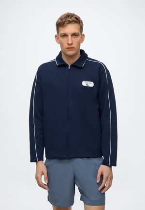 Jeune homme portant une veste zippée bleu marine avec des passepoils blancs et un patch sur la poitrine, assortie à un short bleu clair sur un fond uni.