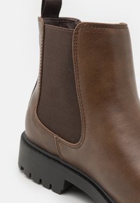 Bota de tobillo de cuero marrón con paneles laterales elásticos, un acabado texturizado y una suela de goma negra gruesa con surcos para un mejor agarre.