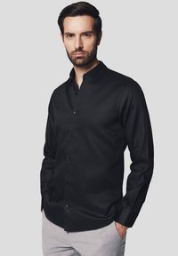 Czarna koszula na długi rękaw z kołnierzykiem, gładką fakturą, guzikami z przodu i dopasowanym krojem. W zestawie z jasnoszarymi spodniami.