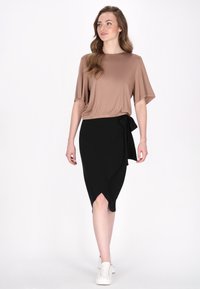 Bruine top met korte mouwen en een gathering in de taille, gecombineerd met een zwarte wikkelrok met een strikaccent. Gedirigeerd met witte sneakers.