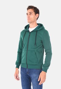 Felpa verde con zip, dotata di tasca frontale, coulisse sul cappuccio e polsini a coste, abbinata a jeans blu. Realizzata in morbido materiale misto cotone.