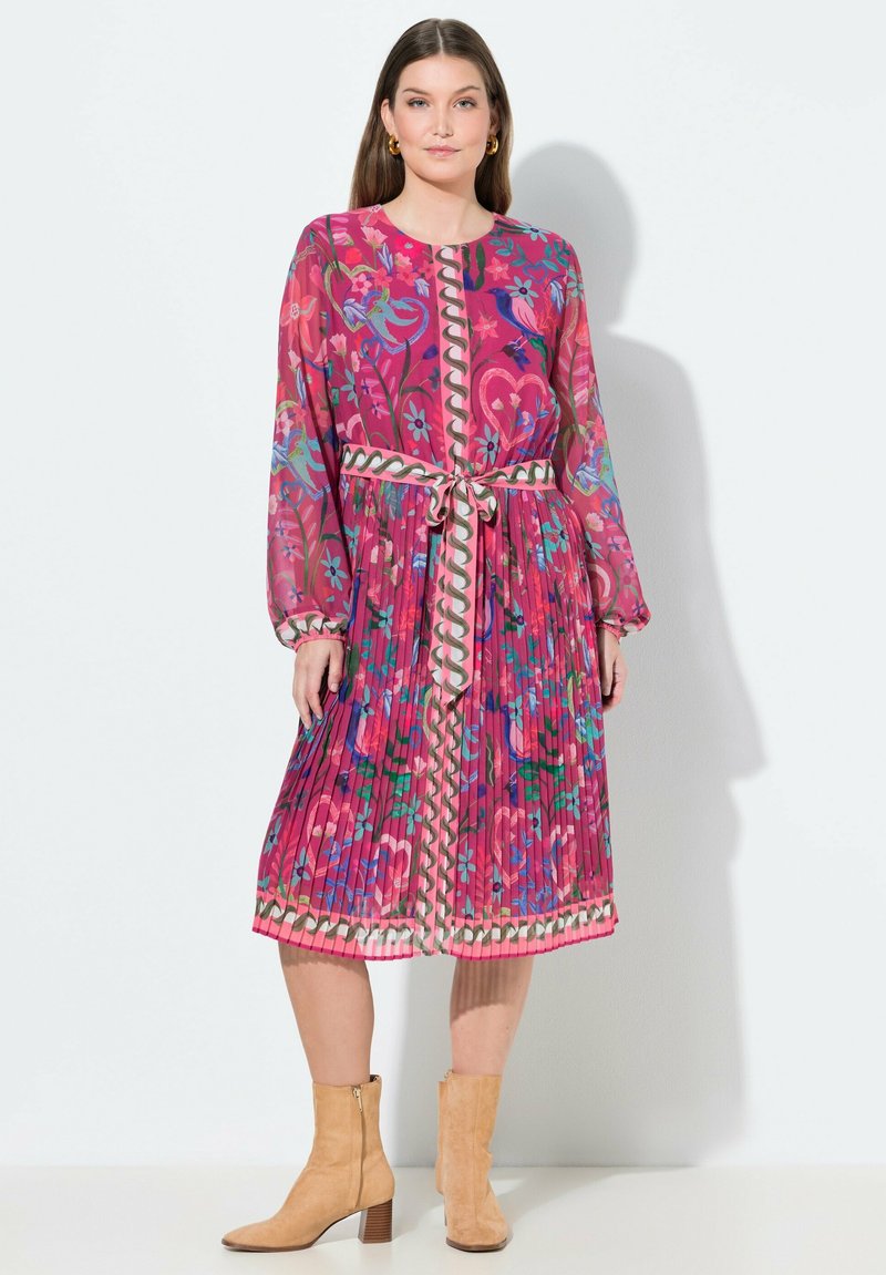 Femme portant une robe midi à manches longues, rose à fleurs avec des plis, une ceinture à la taille et des bottines marron clair, debout devant un fond clair.