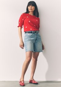 Woman standing wearing a red short-sleeve top with white flowers, light blue denim shorts, black belt, and red pointed flats.