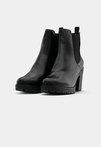 Un par de botas de cuero negro hasta el tobillo con tacones gruesos y paneles laterales elásticos sobre un fondo claro y liso.