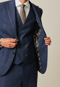 Traje de cuadros azul marino con chaleco de doble botonadura. El interior de la chaqueta presenta un forro con un patrón geométrico. Textura suave, ajuste a medida.